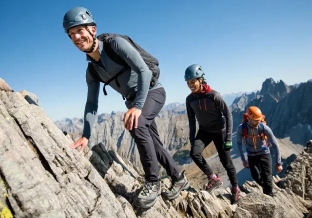 Three alpine scramblers with essential gear ascend a rocky mountain ridge under a clear sky, illustrating non-negotiable equipment for essential gear for alpine scrambling and easy mountaineering.