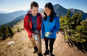 Gaia vs. AllTrails & More: Best Hiking Apps of 2025 Two hikers on a mountain trail comparing different hiking GPS apps on their smartphones, representing the best hiking apps of 2025.
