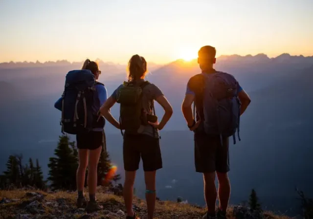 Three diverse backpackers, two women and one man, stand on a mountain ridge at sunrise, overlooking a vast wilderness with their packs, symbolizing the transition from day hiking to backpacking.