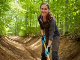 A female hiker performing trail maintenance on a forest path, using a tool to clear a drain as part of a guide to trail stewardship.
