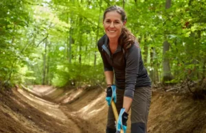 A Hiker’s Guide to Trail Maintenance A female hiker performing trail maintenance on a forest path, using a tool to clear a drain as part of a guide to trail stewardship.