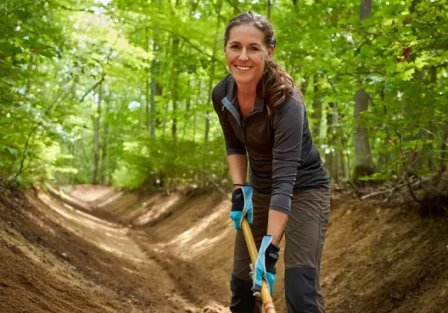 A female hiker performing trail maintenance on a forest path, using a tool to clear a drain as part of a guide to trail stewardship.