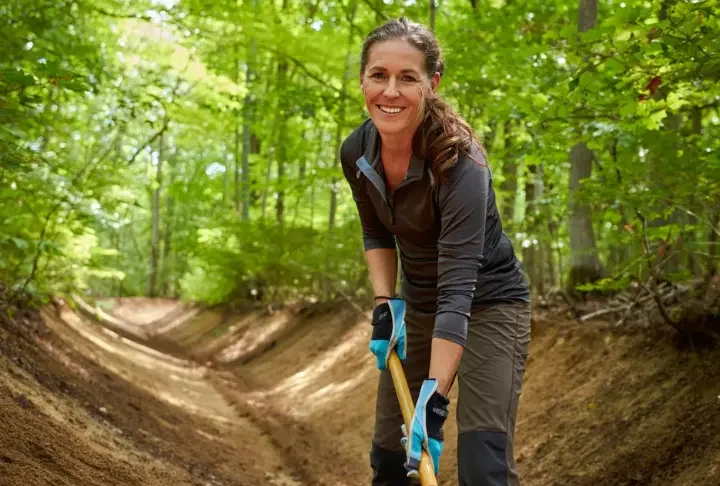 A female hiker performing trail maintenance on a forest path, using a tool to clear a drain as part of a guide to trail stewardship.