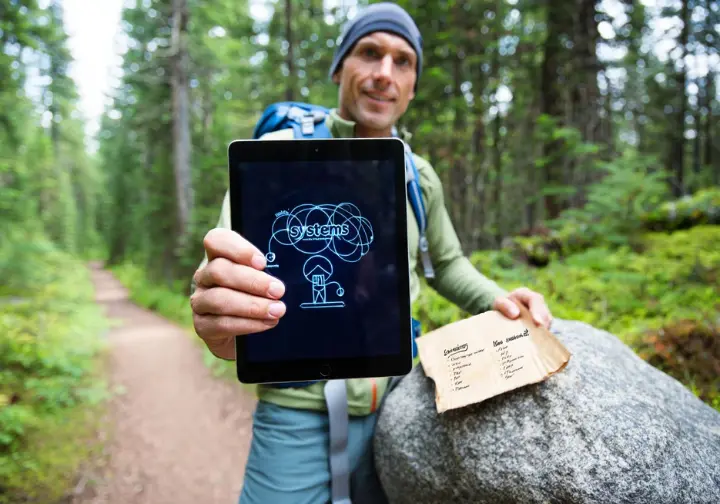 A hiker compares a modern digital "systems" approach to hiking essentials with a traditional paper list, symbolizing the evolution of outdoor preparedness.