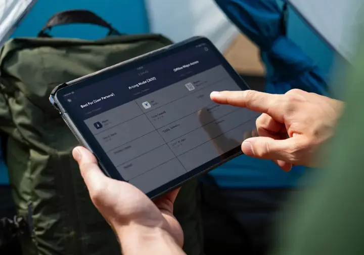Hands interacting with a tablet displaying a hiking app comparison matrix for 2025, showing features and pricing of different best hiking GPS apps.