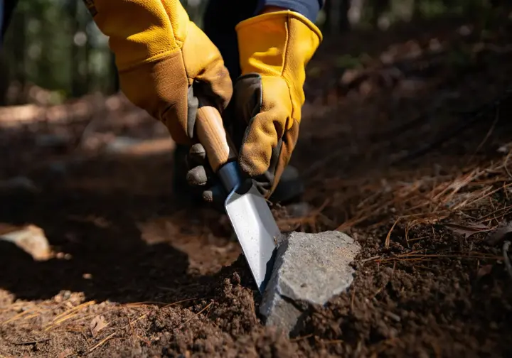 A close-up view of a trail volunteer's gloved hands using a Pulaski tool to do tread work, showing the practice of trail maintenance.
