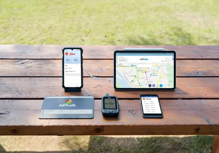 Four digital devices showing different hiking map apps (Gaia GPS, CalTopo, AllTrails, onX Backcountry) laid out, representing a comparative analysis of hiking planning tools.