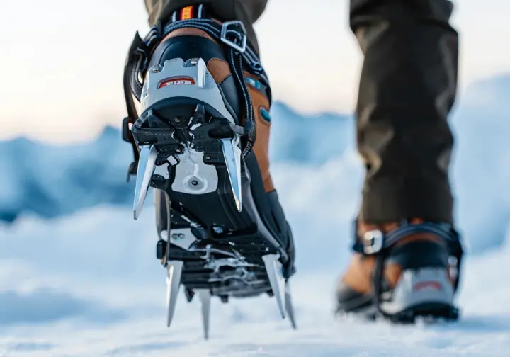 A close-up of hiking crampons with aggressive steel spikes attached to a mountaineering boot, designed for secure movement in mountainous terrain.