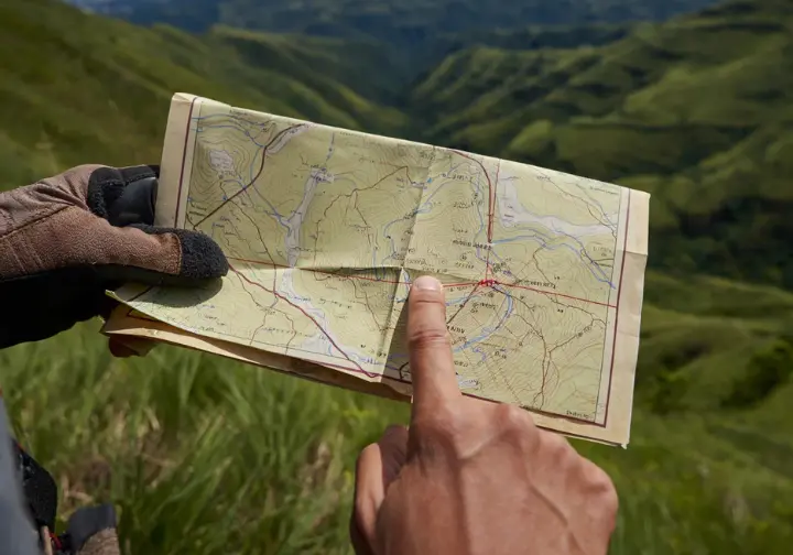 Hands interpreting contour lines on a topographic map with a real-world landscape in the background, illustrating map literacy for navigation.