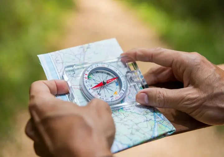 Hands demonstrating how to use a baseplate compass with a topographic map, showing the process of taking a bearing for navigation.