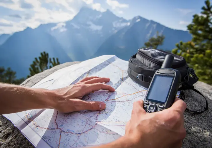 A hiker's hands examine a paper trail map, with a GPS device and a lightweight backpack nearby, illustrating the logistical planning, gear, and trail life of a thru-hiker.