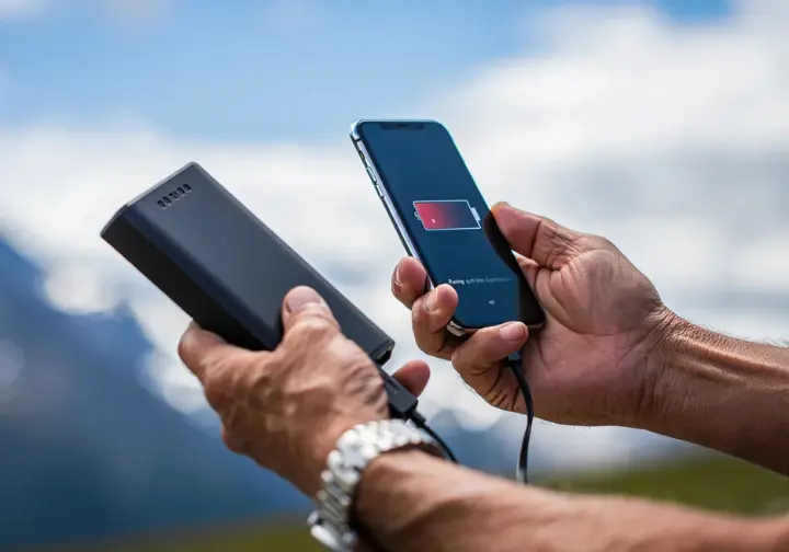 A hiker plugging a power bank into a smartphone displaying a hiking app with a low battery, illustrating solutions to common hiking app problems like battery drain.