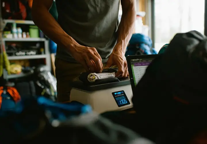 A hiker weighing a backpack on a scale with a digital tablet showing a gear spreadsheet, illustrating practical steps for beginning an ultralight backpacking journey.