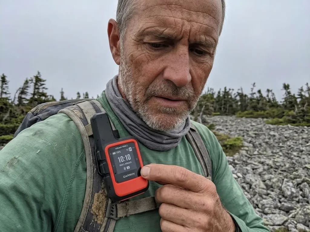 Hiker thumbing a Garmin inReach Mini 2 satellite messenger clipped to a pack strap