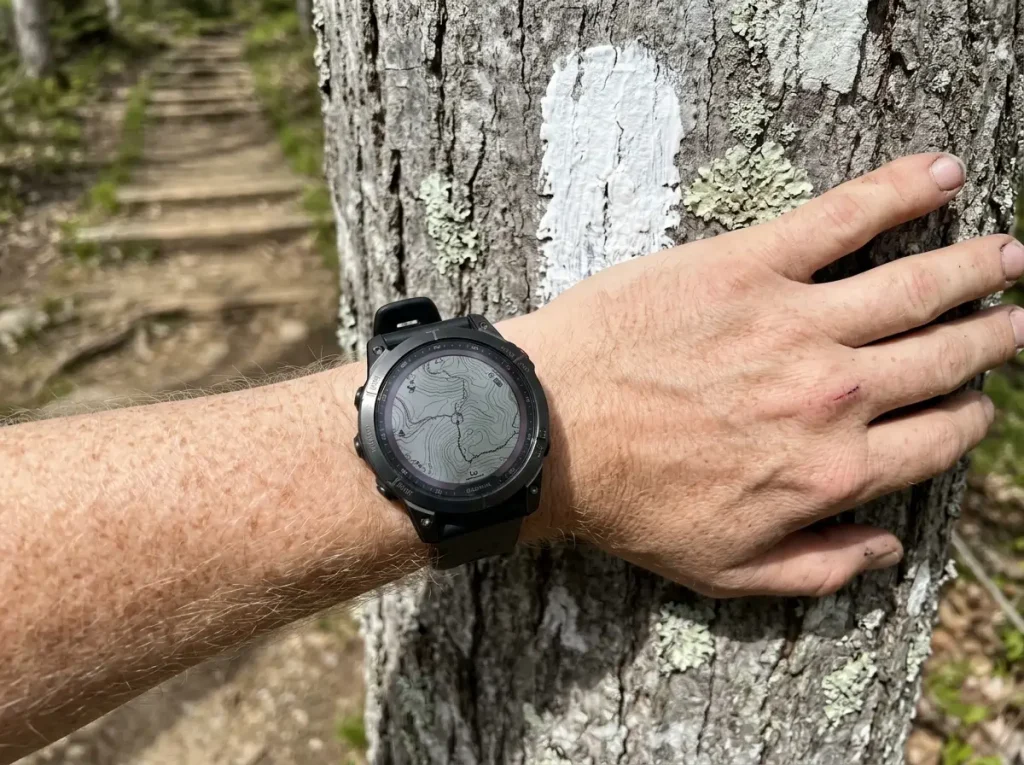 Hiker's wrist GPS watch beside a painted trail blaze on a tree