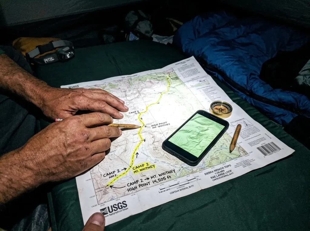 Route planning with a hiking app on a phone next to a paper topo map by headlamp