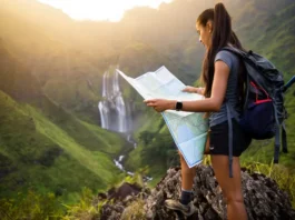 A hiker with a backpack and map plans her route while looking out at a stunning, large waterfall in a mountain valley at sunset.