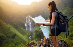 Find a Waterfall Hike: Maps, Gear & Pro Trail Tips A hiker with a backpack and map plans her route while looking out at a stunning, large waterfall in a mountain valley at sunset.