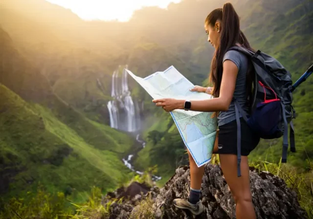 A hiker with a backpack and map plans her route while looking out at a stunning, large waterfall in a mountain valley at sunset.
