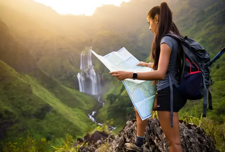 A hiker with a backpack and map plans her route while looking out at a stunning, large waterfall in a mountain valley at sunset.