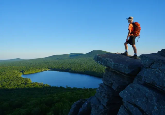 A hiker stands on the rocky summit of Chauncey Peak, looking at the panoramic view of Crescent Lake and Lamentation Mountain in Doctor Francis Giuffrida Park, Meriden, CT.