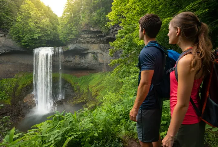 Wide view of the two-tiered Kaaterskill Falls in the Catskills, with two hikers on the viewing platform enjoying the scenic landscape.
