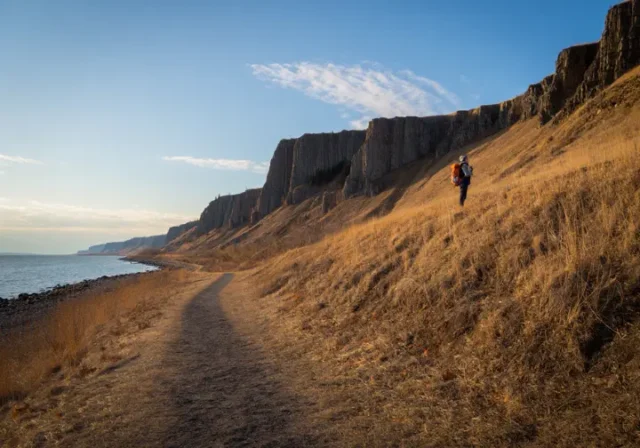 A wide, scenic view of a hiking trail in Palisades Interstate Park at sunset, showing the path along the Hudson River and up the cliffs.