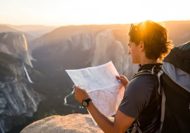 A hiker plans their route with a map while overlooking a vast California mountain landscape during a golden sunset, representing planning hiking trips in California.