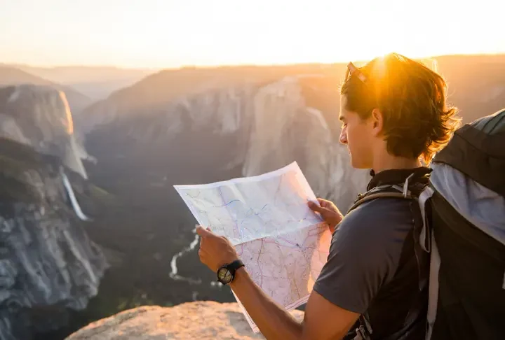 A hiker plans their route with a map while overlooking a vast California mountain landscape during a golden sunset, representing planning hiking trips in California.
