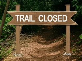 A hiker in full gear stands before a "Trail Closed" sign in a dense forest, looking towards a new trail reroute that branches off to the side.