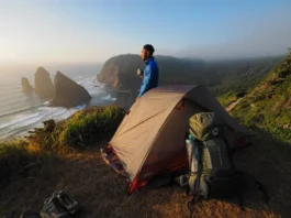 A hiker stands next to their tent at a campsite on a cliff, looking out at a coastal trail, embodying the Vancouver Island camping master plan.