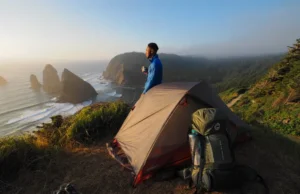 Vancouver Island Camping: The Hiker’s Master Plan A hiker stands next to their tent at a campsite on a cliff, looking out at a coastal trail, embodying the Vancouver Island camping master plan.