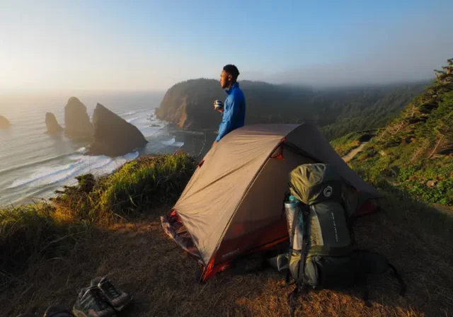 A hiker stands next to their tent at a campsite on a cliff, looking out at a coastal trail, embodying the Vancouver Island camping master plan.