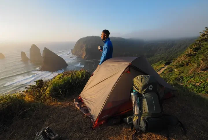 A hiker stands next to their tent at a campsite on a cliff, looking out at a coastal trail, embodying the Vancouver Island camping master plan.