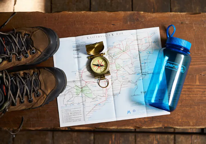 A trail map of Kaaterskill Falls laid out on a wooden table with a compass and hiking boots, representing a trail guide and planning.