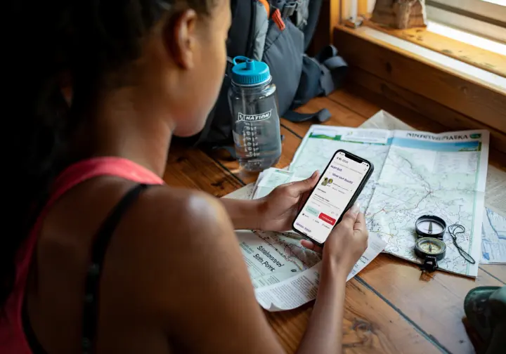 A hiker plans a trip to Sam's Point, using a smartphone to make a reservation online with a trail map and compass on the table.