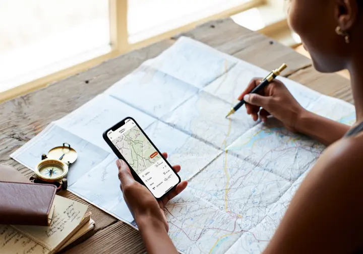 A flat lay of hiking-planning tools on a wooden table, with hands using a smartphone app and a paper topographic map.