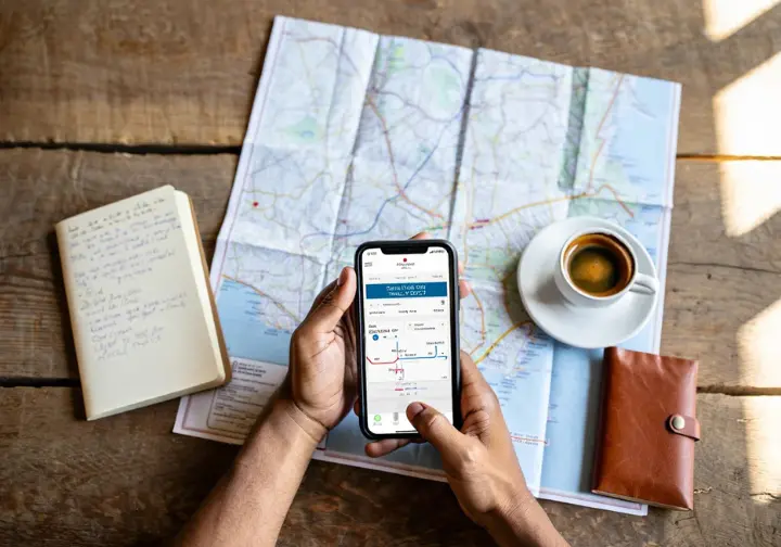 A hiker plans a car-free trip in Europe using a smartphone transport app and a physical trail map on a wooden table.
