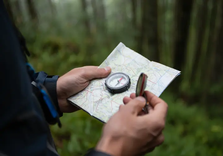 A close-up of a hiker's hands using a map and compass to navigate in a dense forest, demonstrating essential backcountry safety skills.