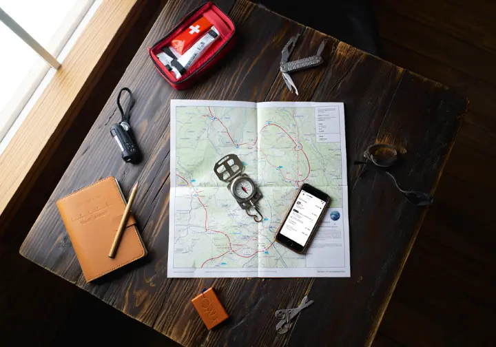 Essential hiking trip-planning toolkit including a map, compass, phone with GPS, and first-aid kit laid out on a wooden table.