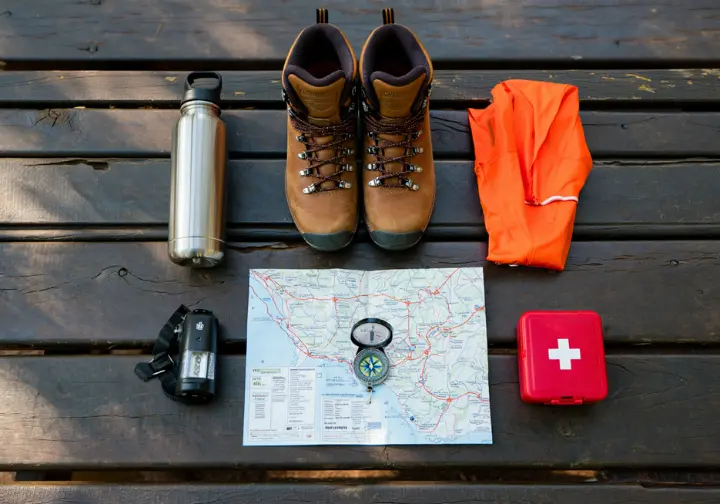 A flat lay of essential hiking gear for a safe hike in Connecticut, including boots, a map, compass, and blaze orange fabric.