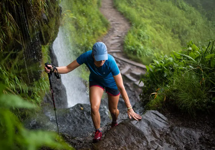 A hiker uses trekking poles for safety and balance while carefully crossing wet, slippery rocks on a trail near a waterfall.
