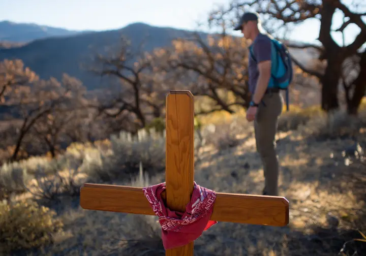 A hiker stands reflectively before a memorial cross on the Storm King Mountain Memorial Trail in Glenwood Springs, Colorado.