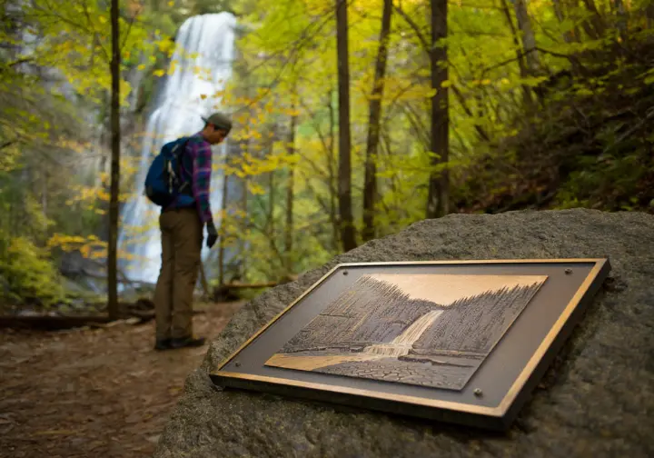 A hiker pauses to read a historical plaque about the Hudson River School, with Kaaterskill Falls in the background, showing the area's deep context.