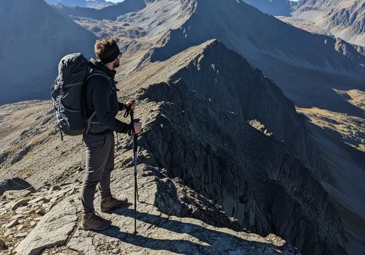 A full-body shot of a lone hiker with a backpack standing on a mountain ridge, looking out over a vast and remote wilderness.