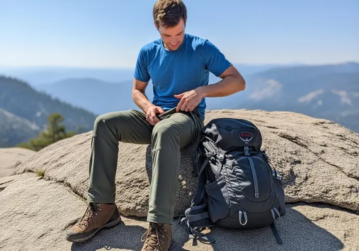 A full-body view of a male hiker sitting on a rock and pointing to the gusseted crotch of his hiking pants.