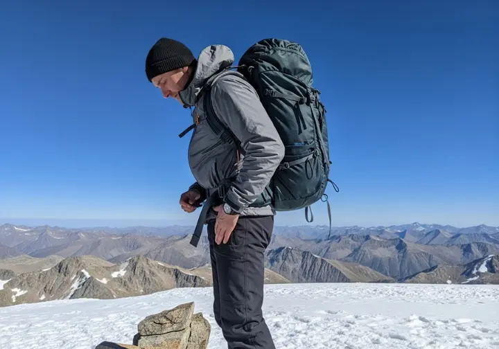 Full-body side view of a hiker on a mountain ridge adjusting the suspension straps of their technical backpack for a better fit