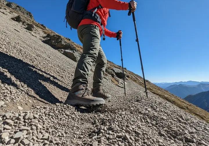 A full-body action shot of a hiker descending a steep scree slope, with the boot's outsole providing critical grip on the loose ground.