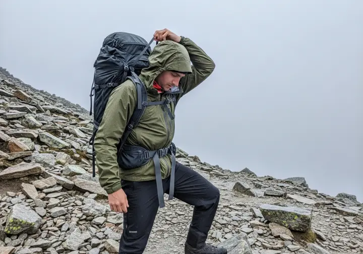 A hiker in full rain gear stops on a trail to make a small adjustment to their backpack's load lifter strap.