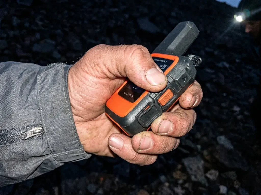 Hiker lifting the SOS cover on a Garmin inReach Mini 2 at dusk under a headlamp